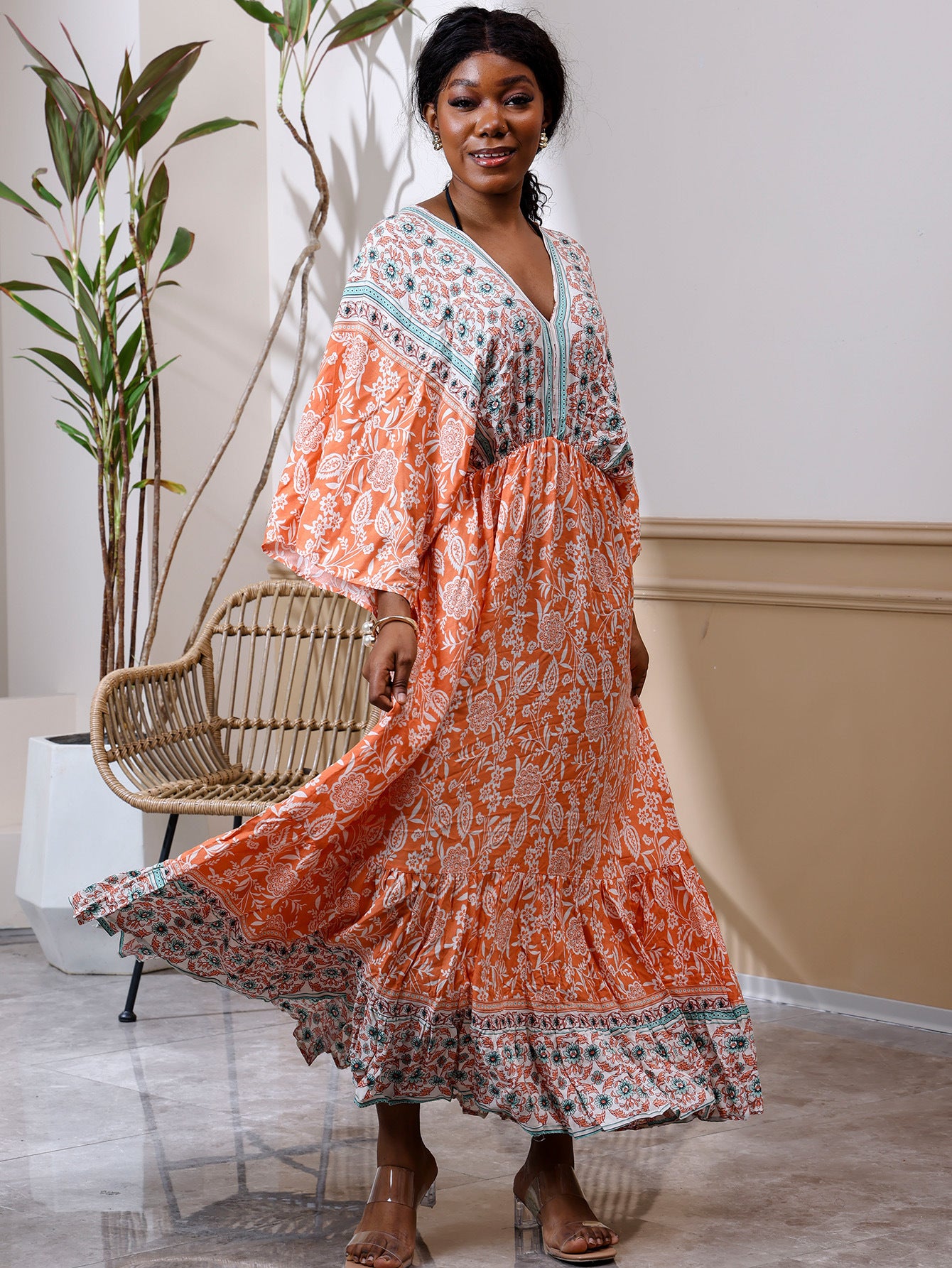 Woman in a long, patterned dress standing in a room with a plant and chair.