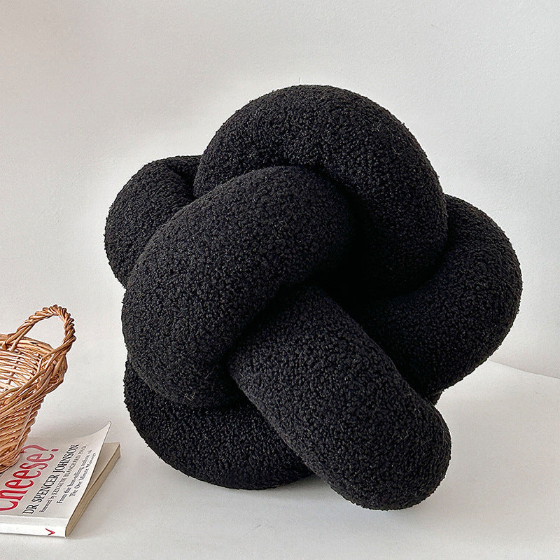 Black knot-shaped pillow on a white surface with a wicker basket and book in the background.