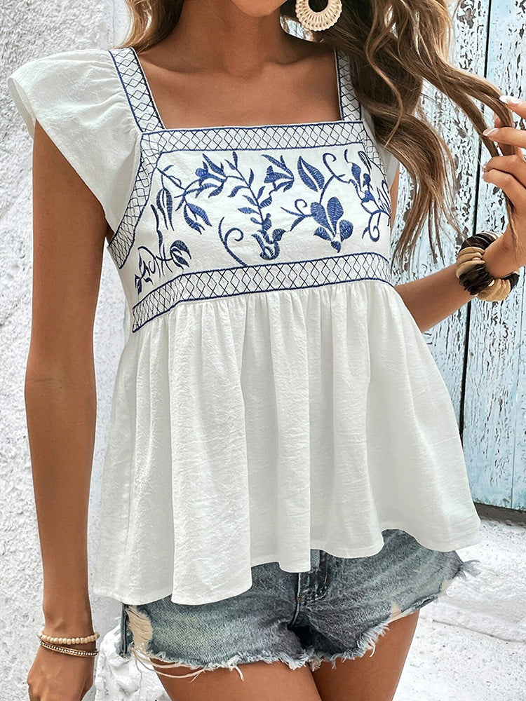 Woman wearing a white top with blue floral embroidery and denim shorts against a light background