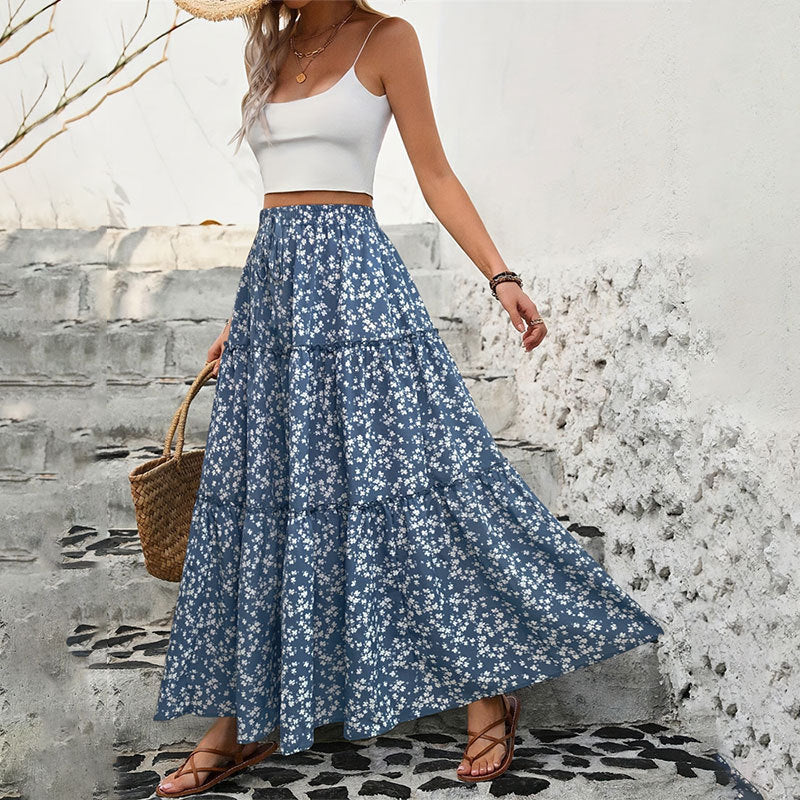 Woman wearing a white top and blue floral skirt with a straw bag against a textured wall.