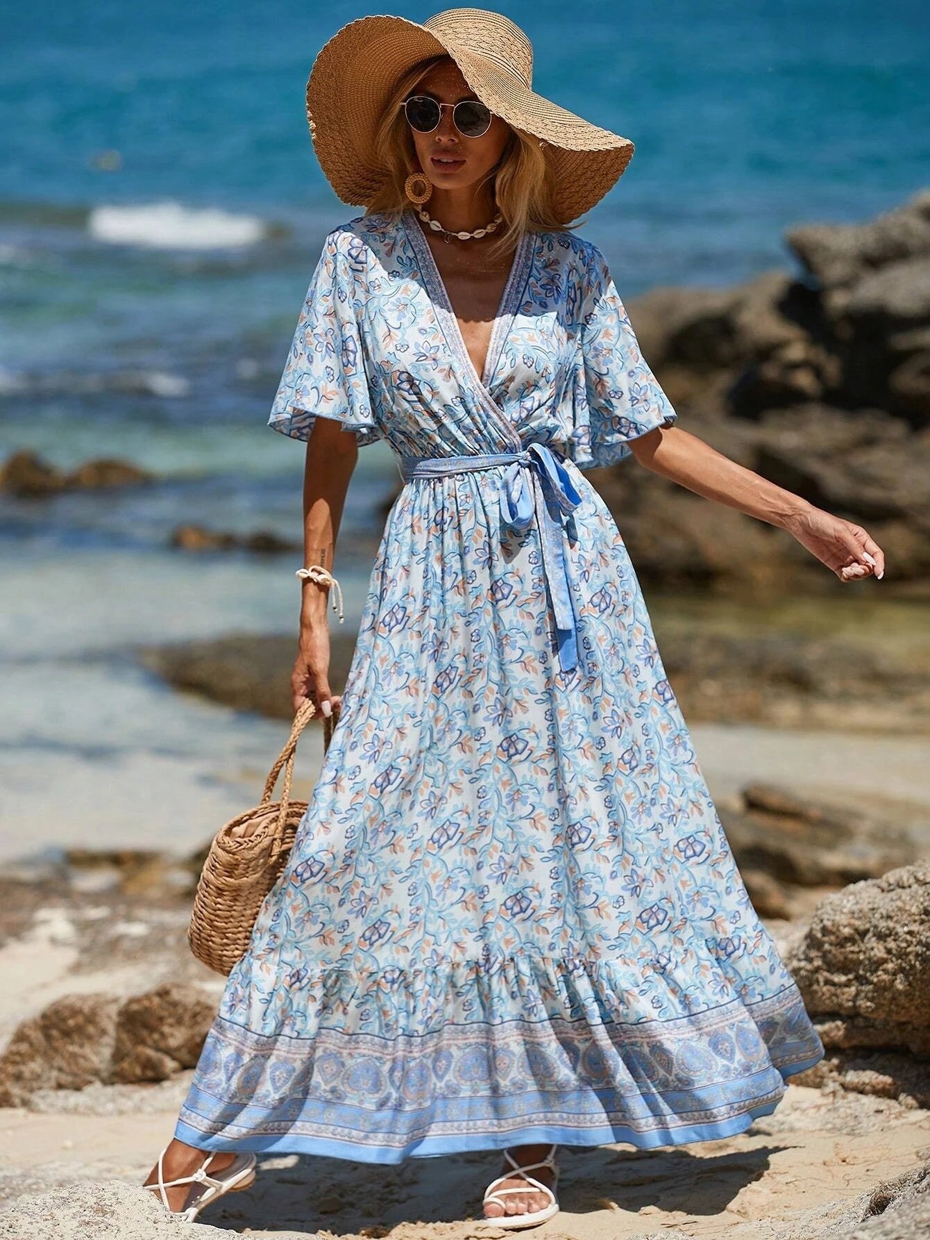 Woman in a blue floral dress and sun hat standing on a rocky beach.
