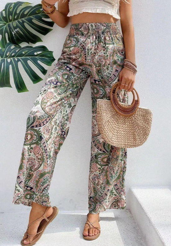 Person wearing floral pants and holding a straw bag against a white background with a leafy plant.