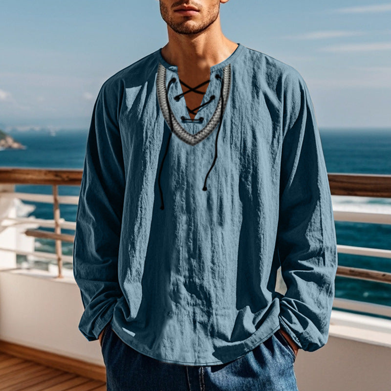 Man wearing a blue long-sleeve shirt with a necklace, standing by a railing with ocean view.