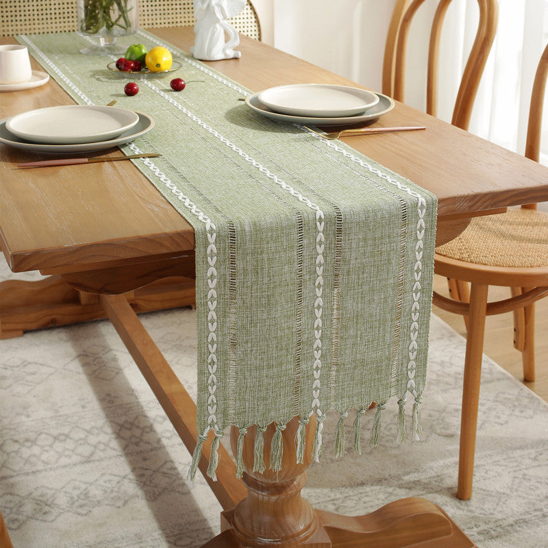 Green table runner with fringe on a wooden dining table with plates and cutlery.