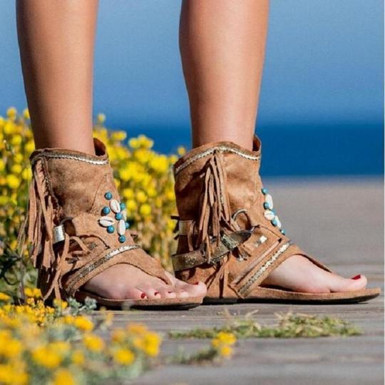 Brown fringe sandals with decorative elements worn by a person standing on a wooden surface with yellow flowers and blue sky in the background.