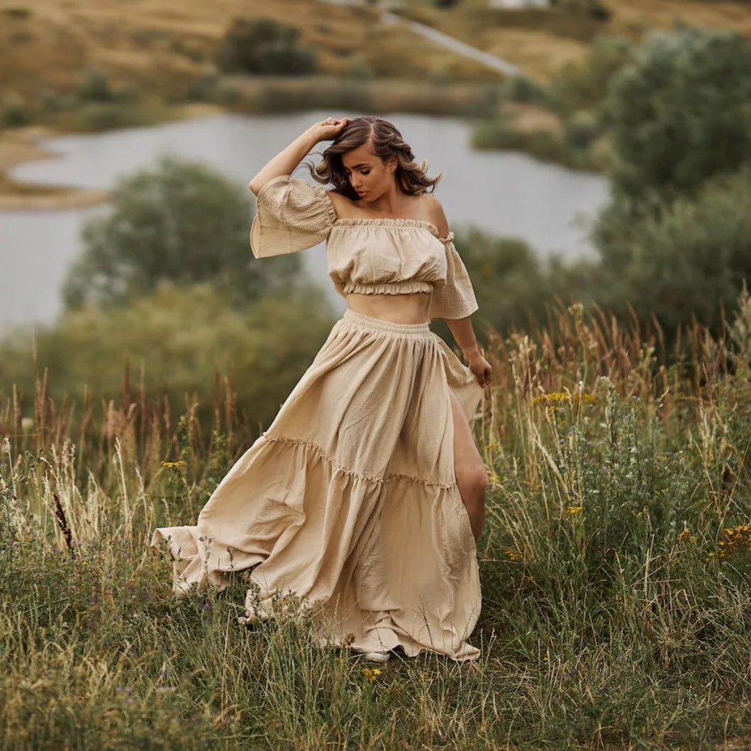 Woman in a flowing beige dress standing in a natural setting with water and greenery.