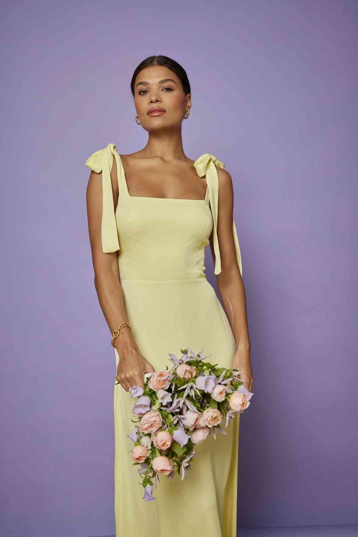 Woman in a light yellow dress with bow straps holding a bouquet against a purple background