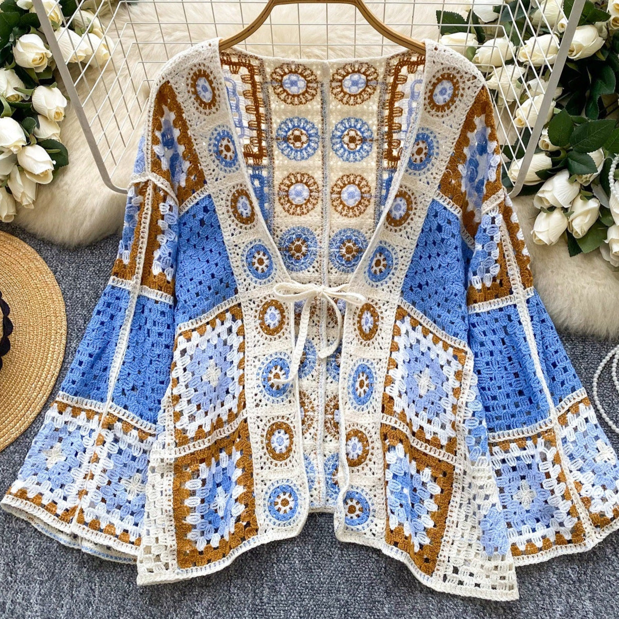 Crocheted cardigan with blue, brown, and white pattern on a hanger with flowers and a hat in the background.