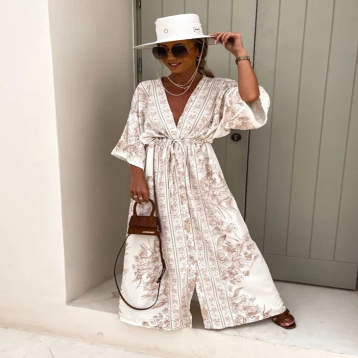 Woman in a white floral dress and hat standing against a neutral wall.