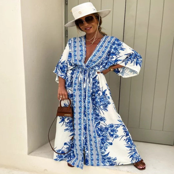 Woman wearing a blue and white floral dress with a white hat and sunglasses, standing against a light-colored wall.