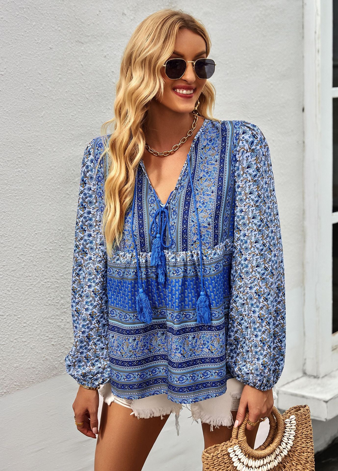 Woman wearing a blue patterned blouse with sunglasses and a handbag, standing against a white wall.