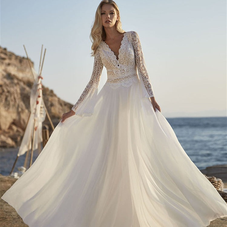 Woman in a white wedding dress with long sleeves standing by the ocean.