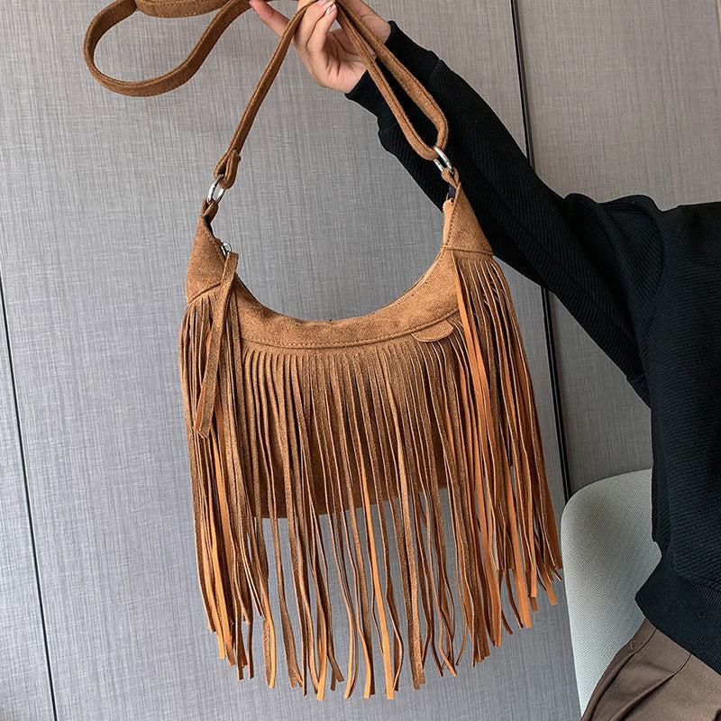 Brown suede handbag with fringe held by a person against a neutral background