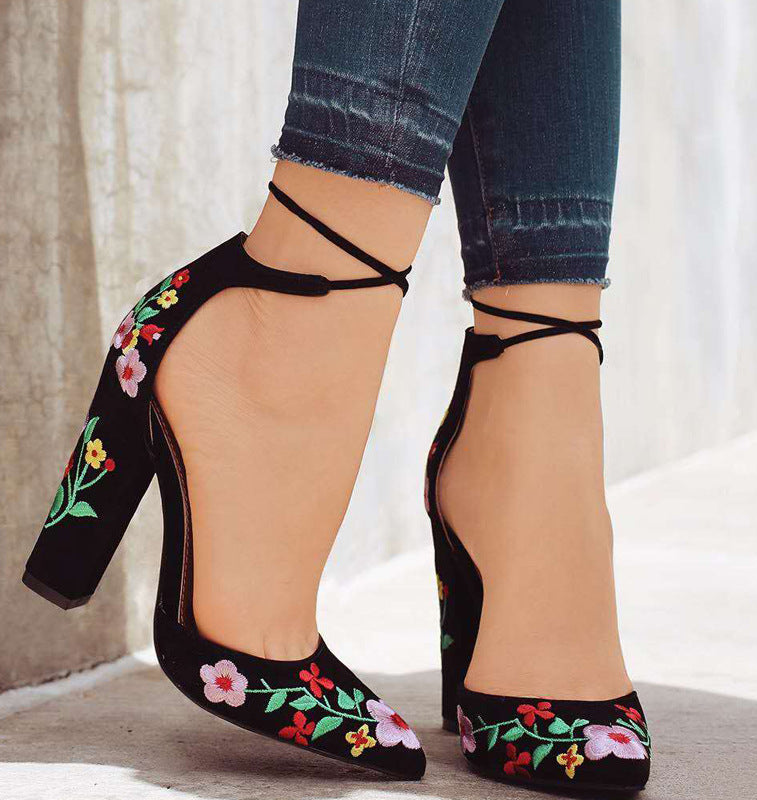 Black high-heeled shoes with floral embroidery worn with blue jeans on a neutral background