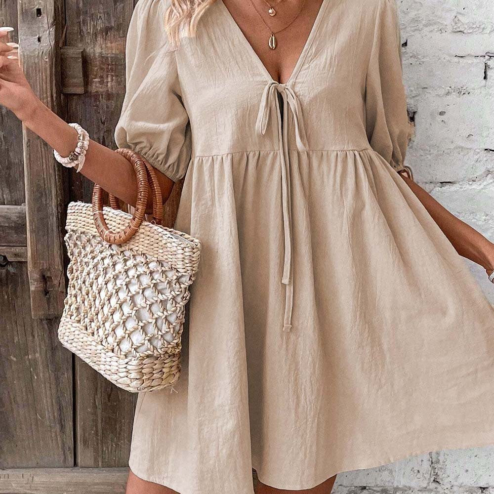 Woman wearing a beige dress holding a woven bag against a rustic wooden and stone wall.