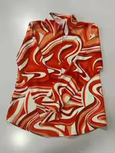 A short-sleeve button-up shirt with a bold abstract swirl pattern in red, orange, and white, laid flat on a light gray background.