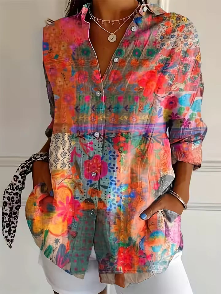 A woman wearing a colorful, long-sleeve button-up shirt with a vibrant floral and abstract patchwork print in shades of pink, orange, green, and blue. The shirt features a split design with a solid floral pattern on the upper chest and a patterned waistband, paired with white shorts.