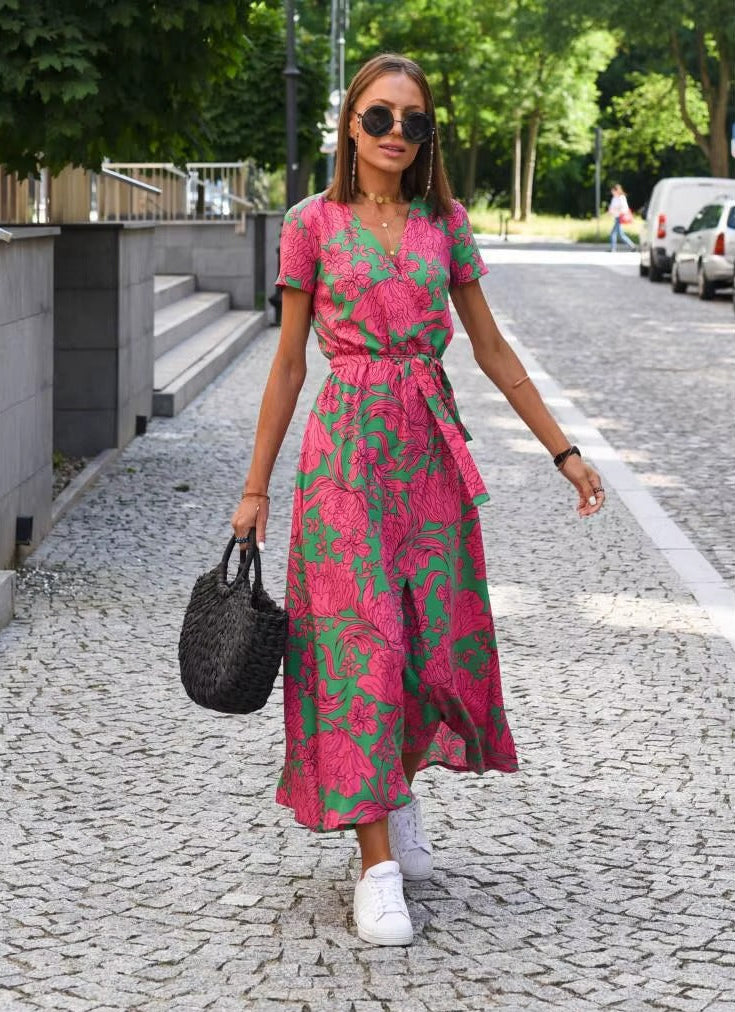 A woman walking on a cobblestone street wearing a pink and green floral print midi dress with short sleeves and a tie waist, holding a black woven handbag.