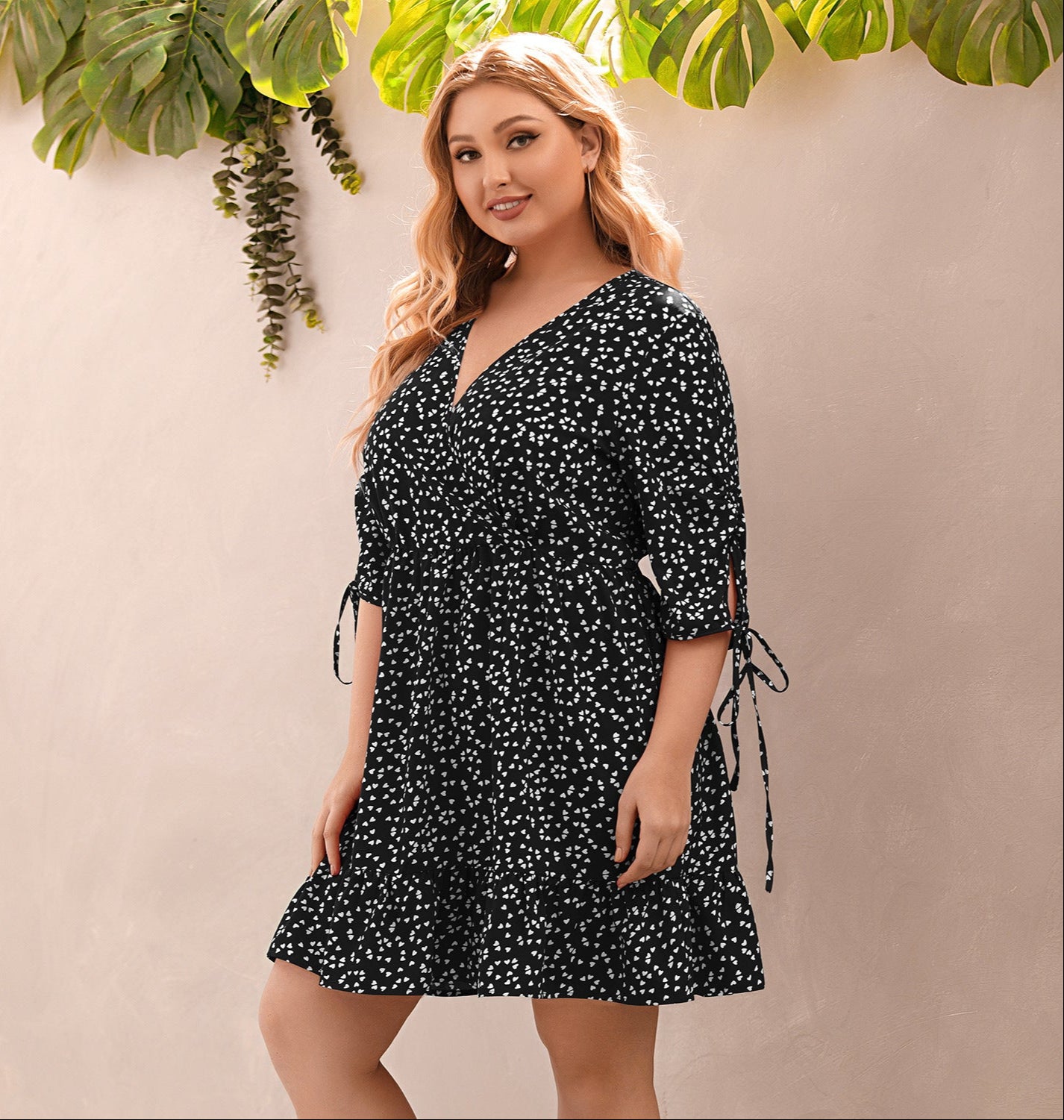Woman wearing a black floral dress against a beige wall with green plants.
