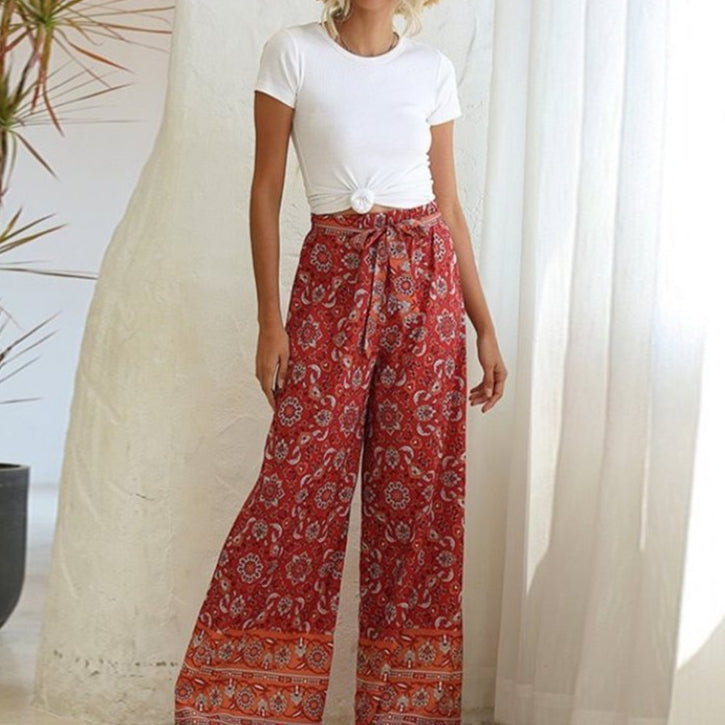 Woman wearing a white top and red patterned pants standing indoors with a plant and curtain in the background.