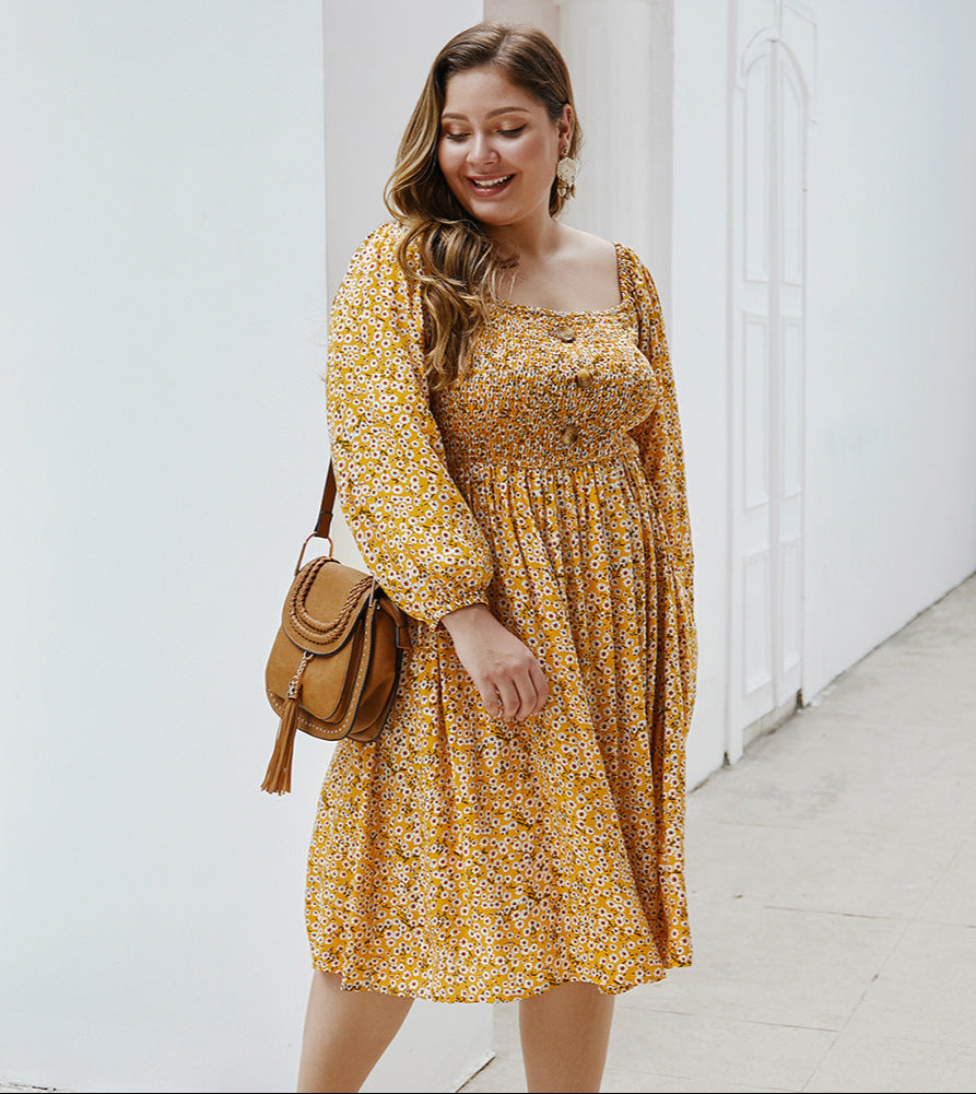 Woman wearing a yellow floral dress with a brown bag, standing against a white wall.