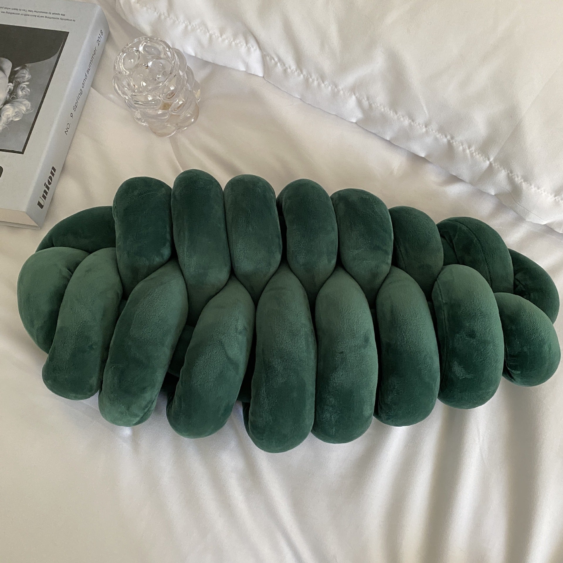 Green plush toy resembling a snake on a white surface with a book and glass object in the background.