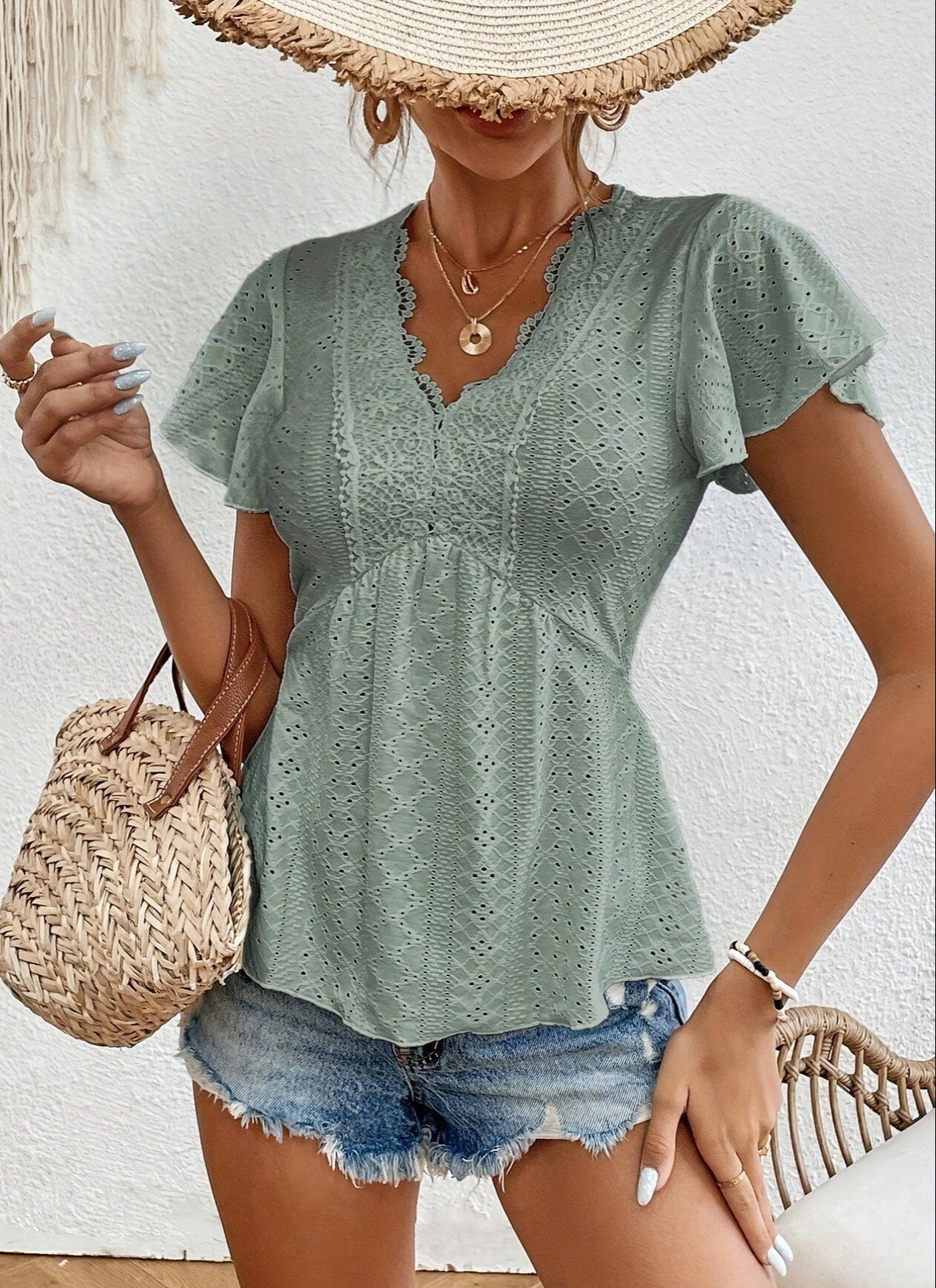 Woman wearing a light green lace top and denim shorts, holding a straw bag against a white wall.