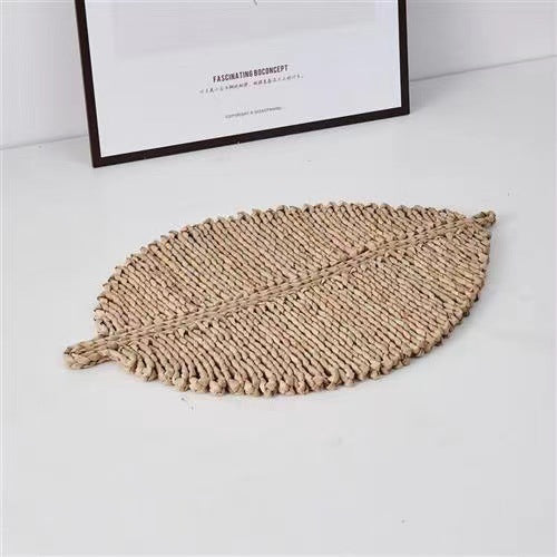 Leaf-shaped woven mat on a white surface with a framed picture in the background.