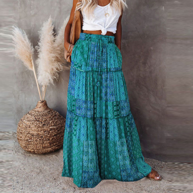 Woman in a white top and green patterned skirt standing next to decorative pampas grass against a neutral wall.