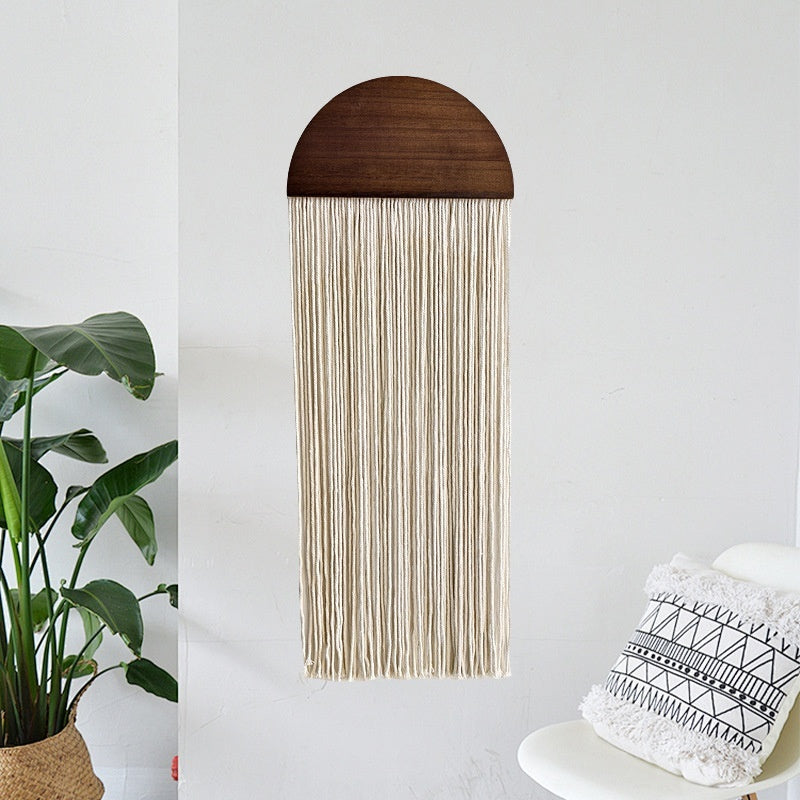 Woven wall art with wooden top section and white strings, placed on a white surface with a plant and pillow in the background.