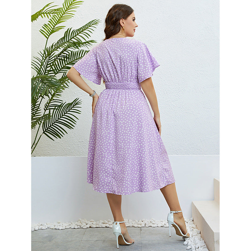 Woman wearing a purple polka dot dress standing against a white wall with a plant.