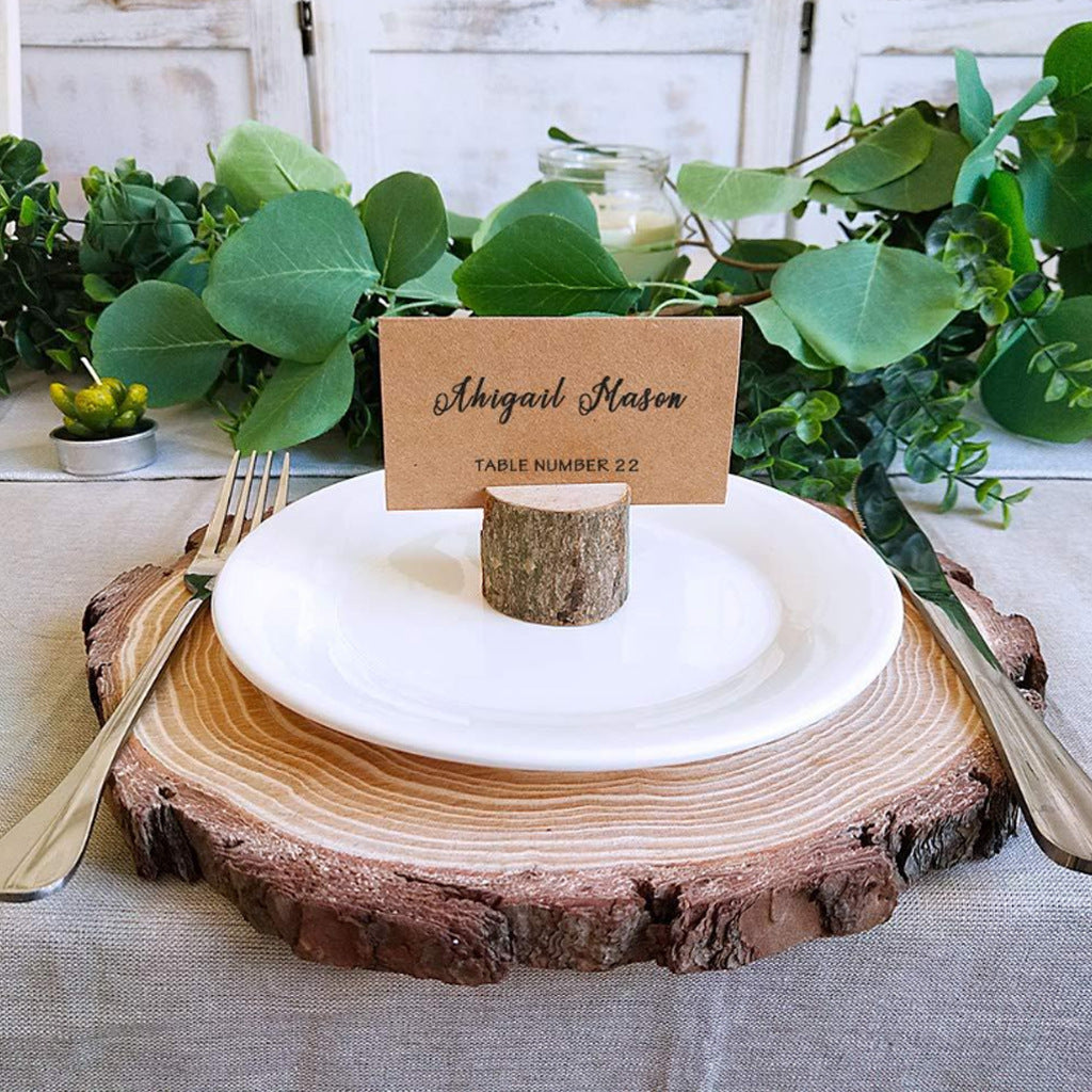Table setting with wooden placemat, white plate, and name card holder on a table with greenery.