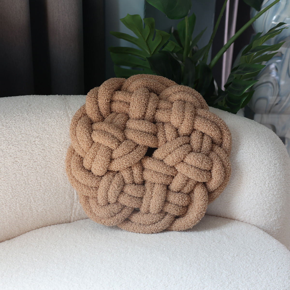 Round braided pillow on a beige sofa with plants in the background