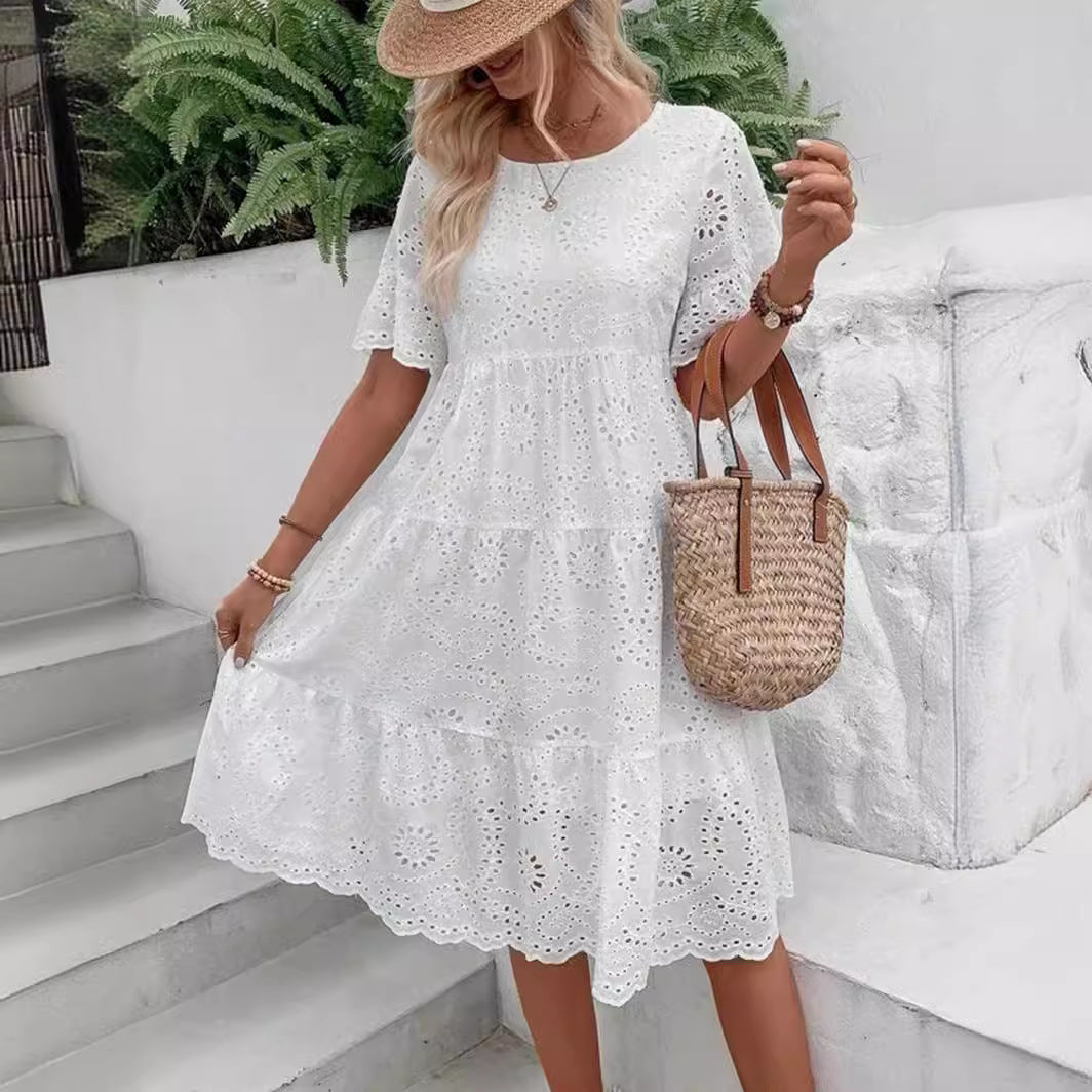 Woman wearing a white lace dress and straw hat, standing on steps with a white wall and greenery in the background.