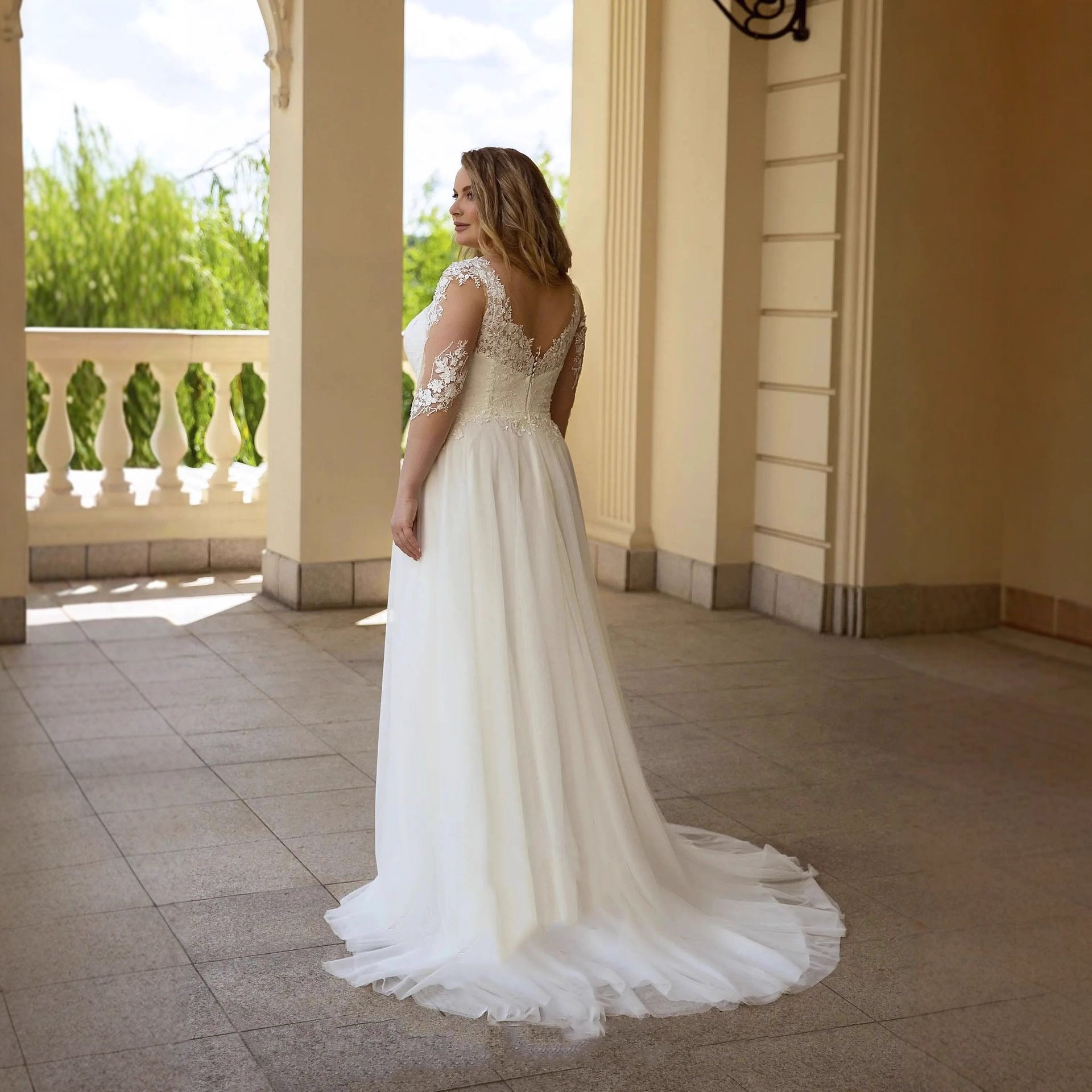 Woman in a white wedding dress standing on a balcony with columns and greenery.