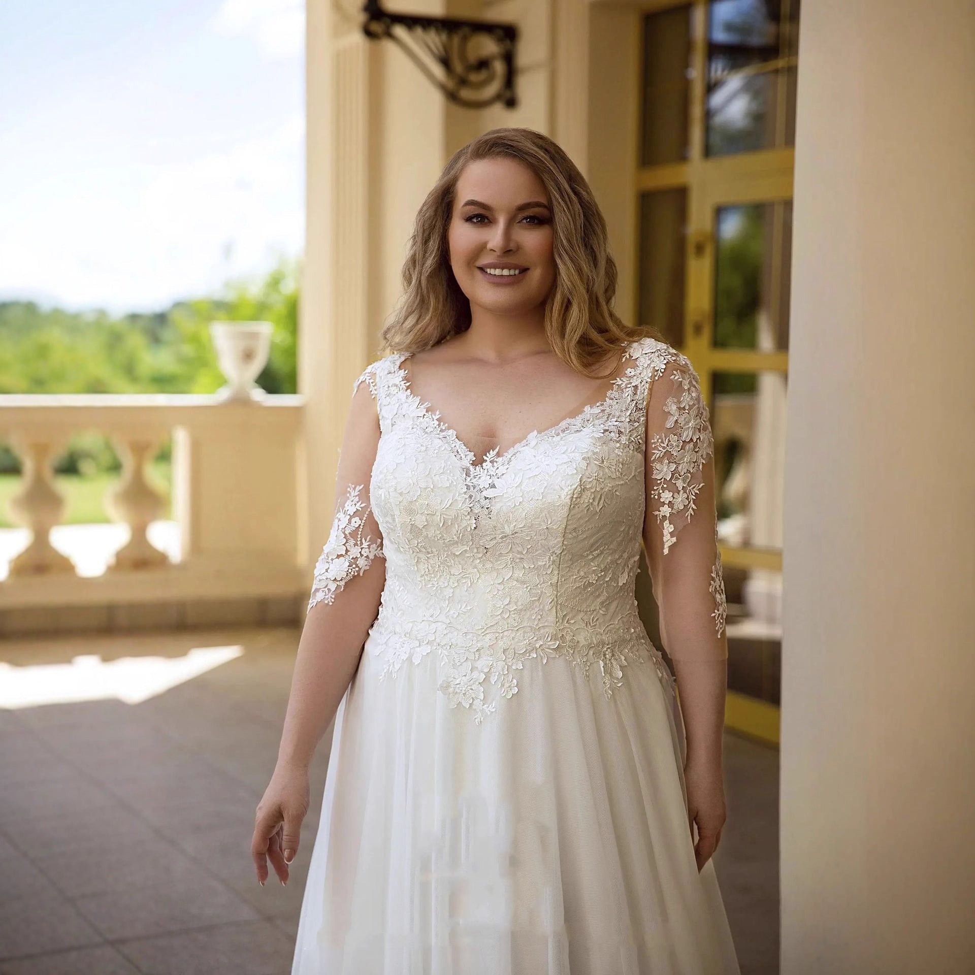 Woman wearing a white lace wedding dress on a balcony with greenery in the background