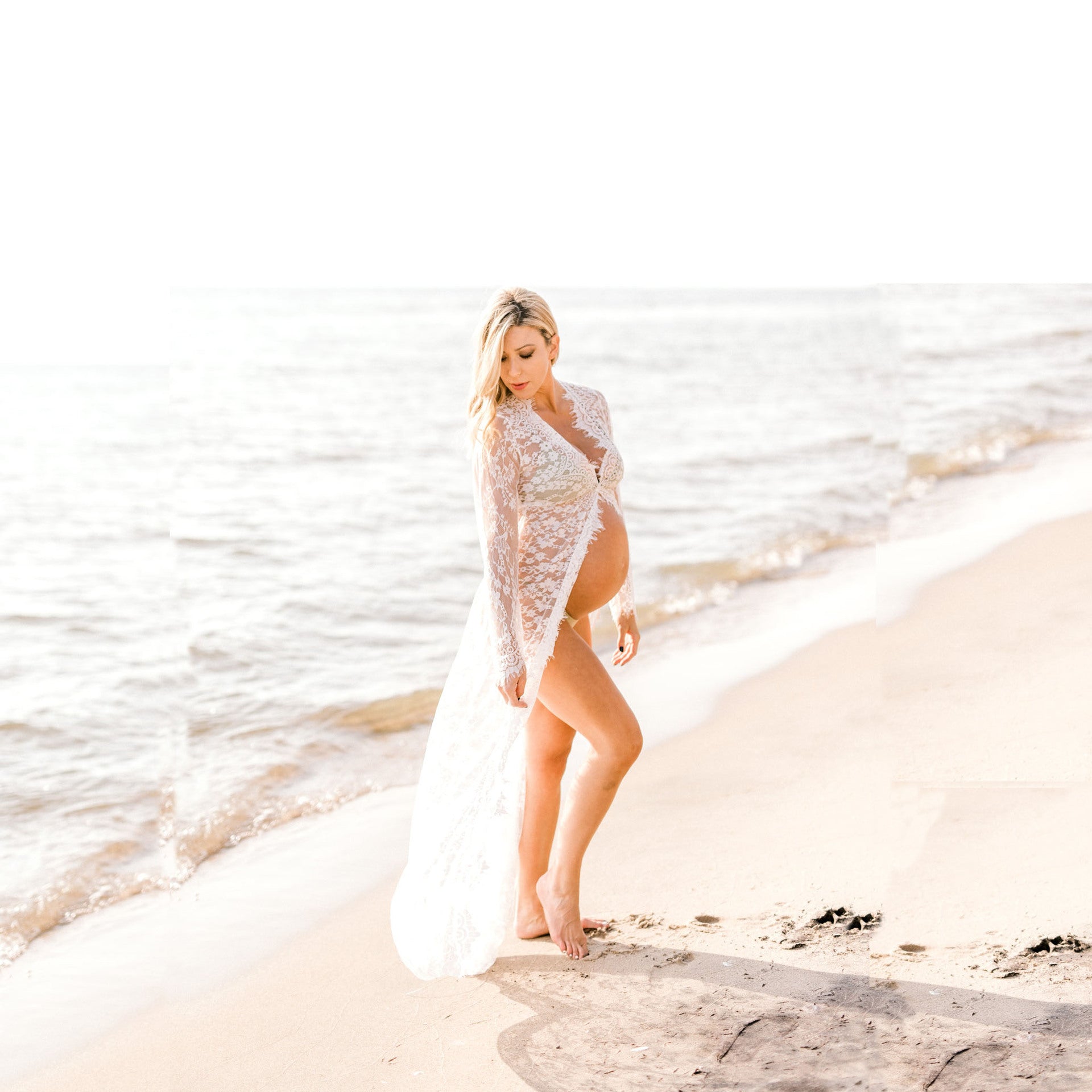 pregnant Woman in a white lace dress standing on a beach with ocean waves in the background