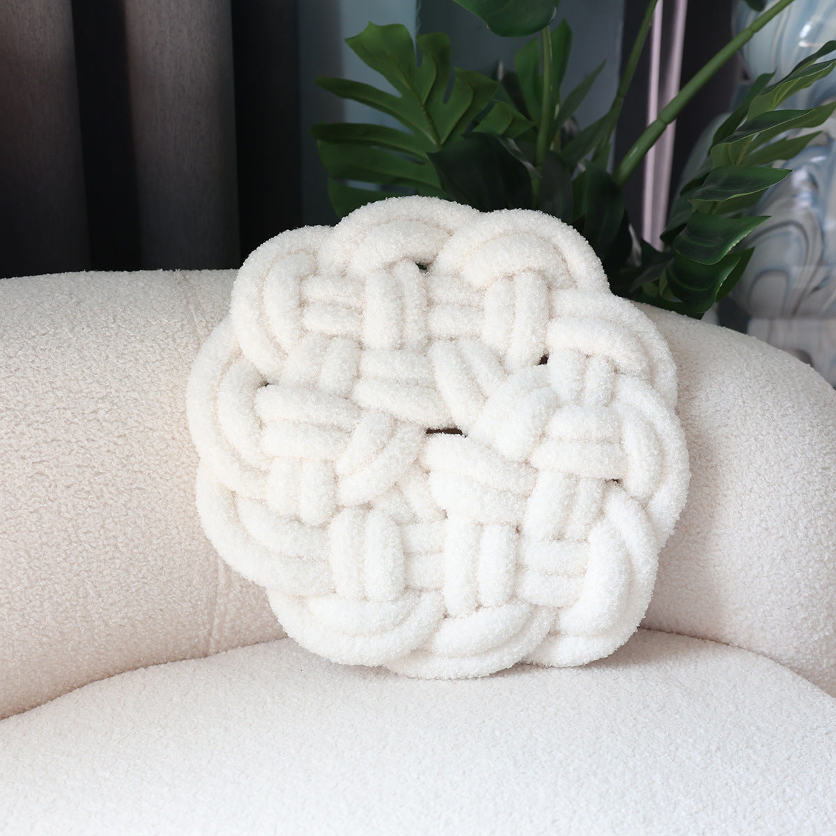 White braided knot pillow on a beige sofa with a plant in the background
