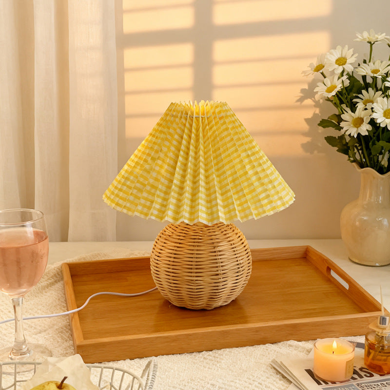 Wicker table lamp with yellow checkered shade on a wooden tray, next to a glass of pink wine and a vase of flowers.