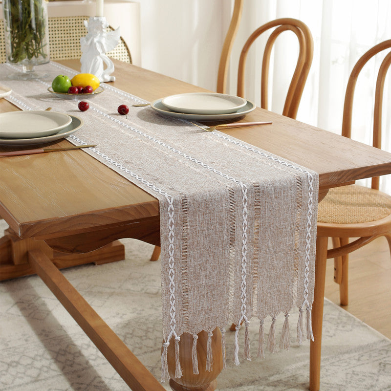 Wooden dining table with a beige table runner and plates on a light-colored floor.