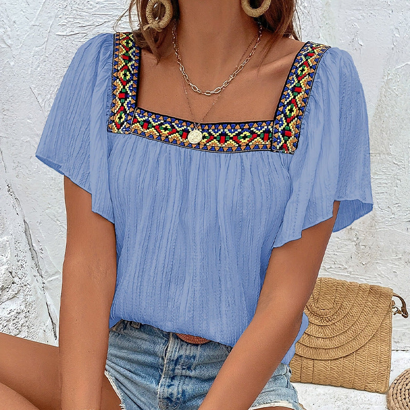Woman wearing a blue top with colorful embroidery, denim shorts, and a straw bag against a light background.