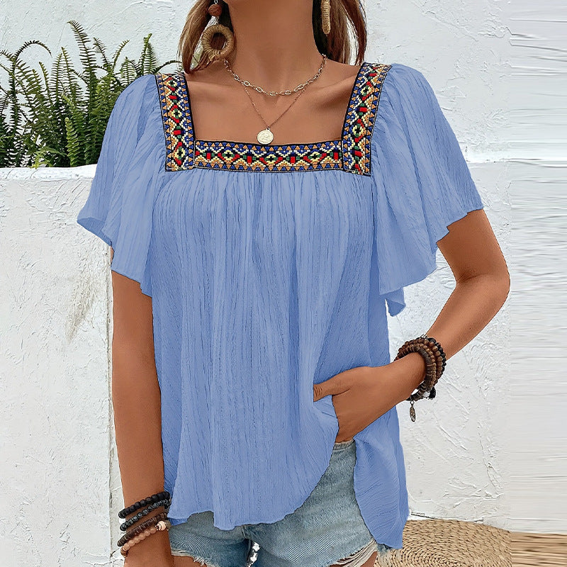 Woman wearing a blue blouse with colorful embroidery against a white wall.