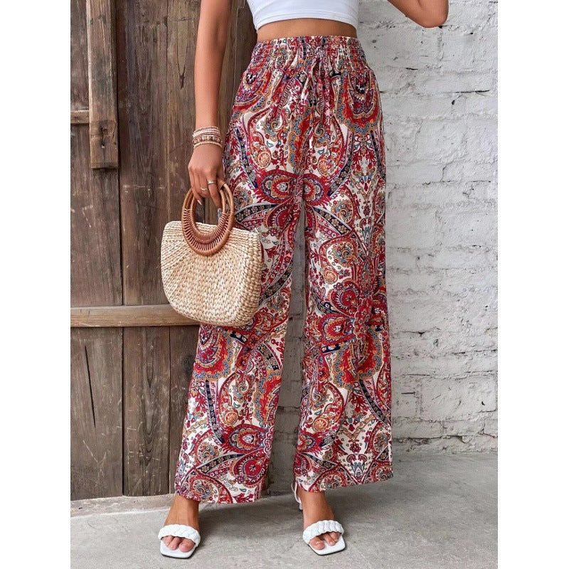 Person wearing paisley patterned pants holding a straw bag against a wooden and stone wall.