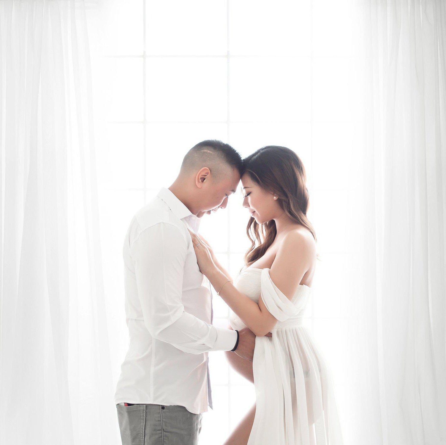 Couple in white clothing standing close together against a white curtain background