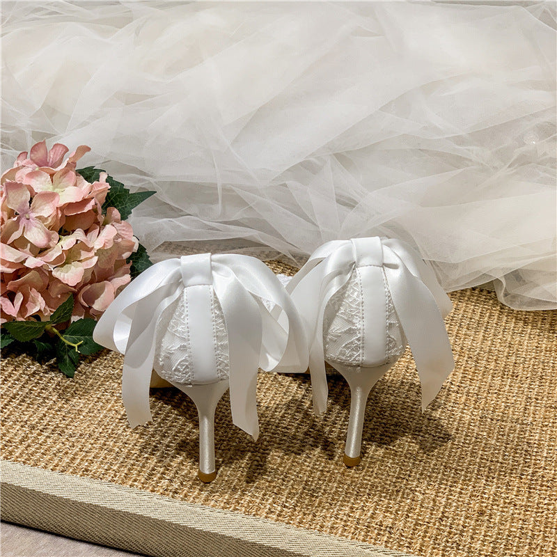 White high-heeled shoes with large bows on a textured surface with flowers and a white fabric background.