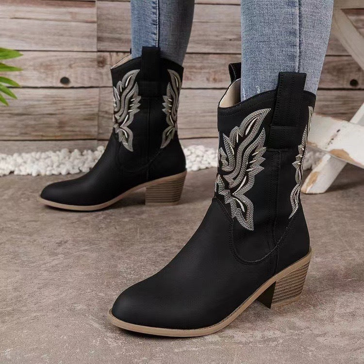 Black cowboy boots with decorative patterns worn by a person on a wooden floor.