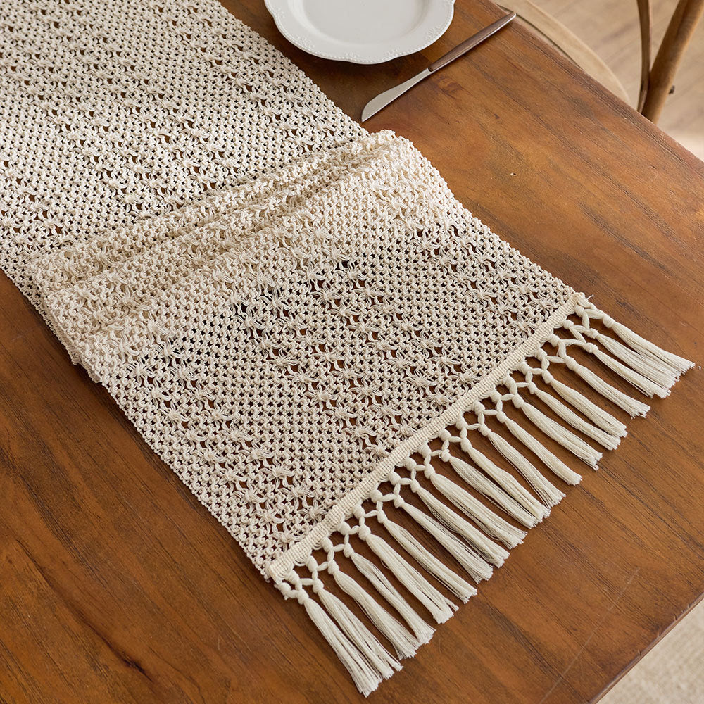 Beige crocheted table runner with tassels on a wooden table.