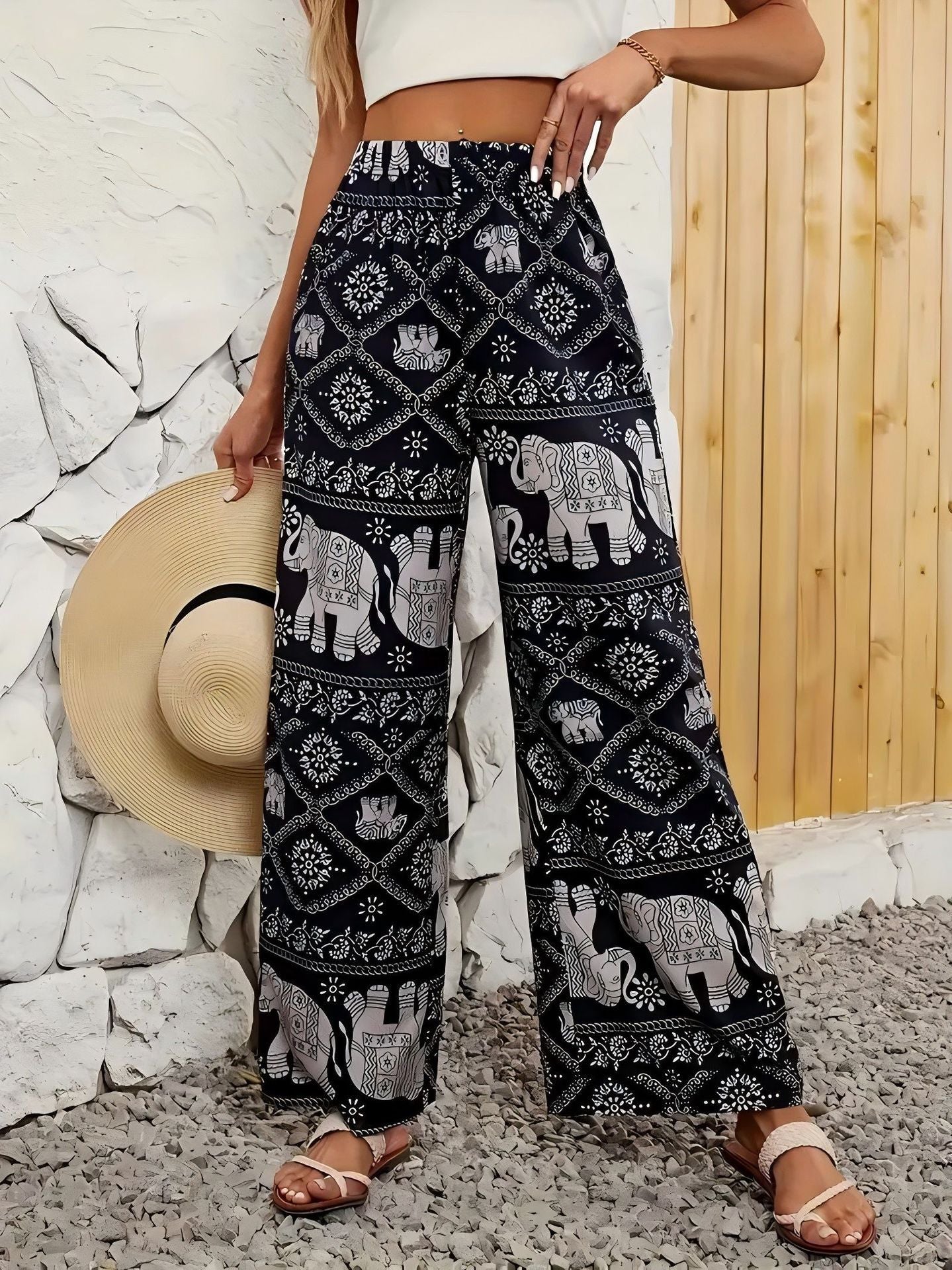 Person wearing black patterned pants with elephants, holding a hat against a stone wall and wooden panel background.