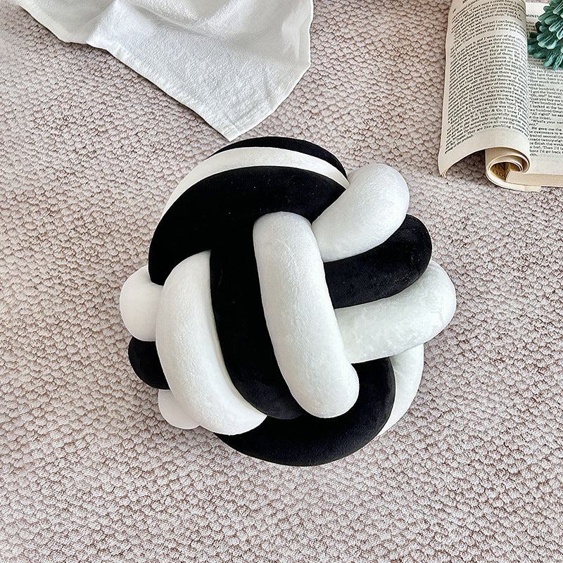 Black and white knot-shaped pillow on a textured surface with a book and towel in the background