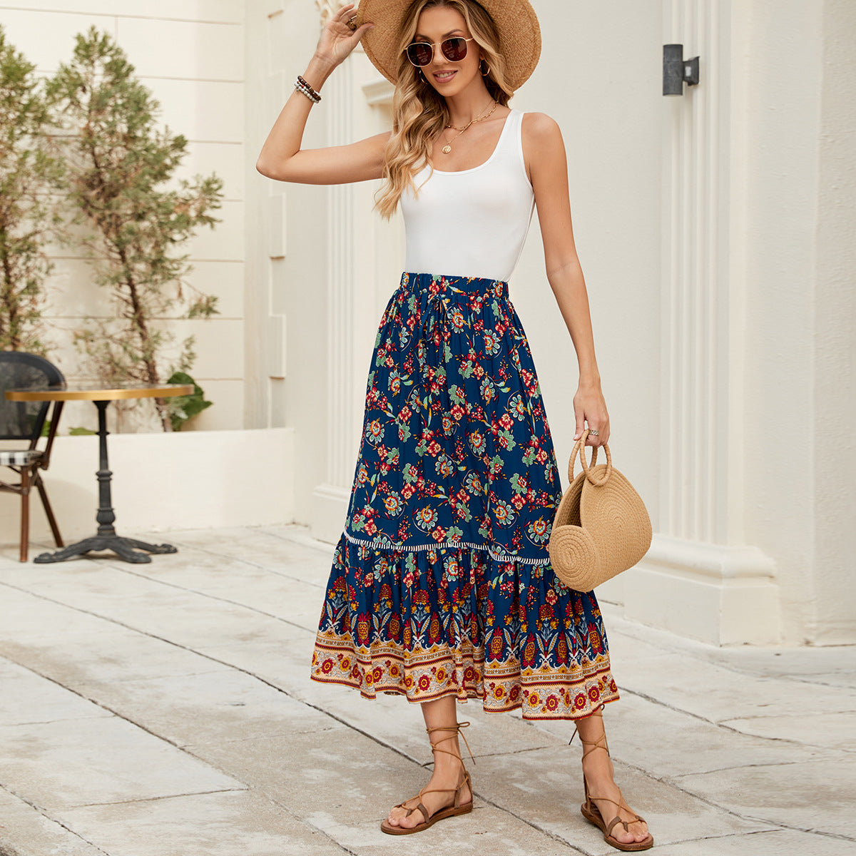 Woman wearing a white top and blue floral skirt with a straw hat and bag, standing on a wooden floor.